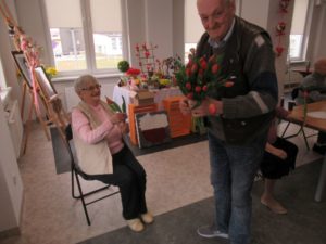 Senior dziennego domu wręcza Seniorce tulipana z okazji Dnia Kobiet, która siedzi na krześle