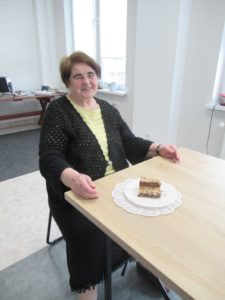 Seniorka uśmiecha się siedząc przy stole, na którym znajduje się ciasto