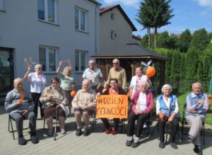 nominowani seniorzy dziennego domu prezentują na zewnątrz budynku pomarańczowe rekwizyty #challengedziengodnosci