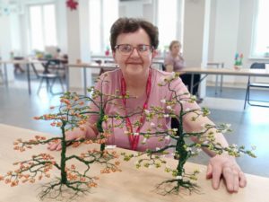 Uśmiechnięta seniorka Dziennego Domu Pomocy siedzi przy stole w Dziennym Domu i prezentuje trzy Drzewka Szczęścia