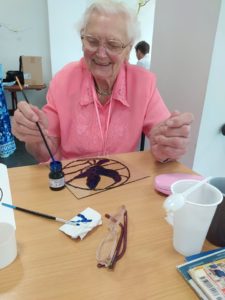 seniorka Dziennego Domu wypełnia farbą kontury pracy na szkle na zajęciach arteterapii