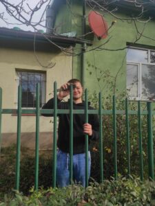 Młody, uśmiechnięty mężczyzna czyści metalowe ogrodzenie . Za nim krzew oraz dwa połączone ze sobą budynki z oknami.